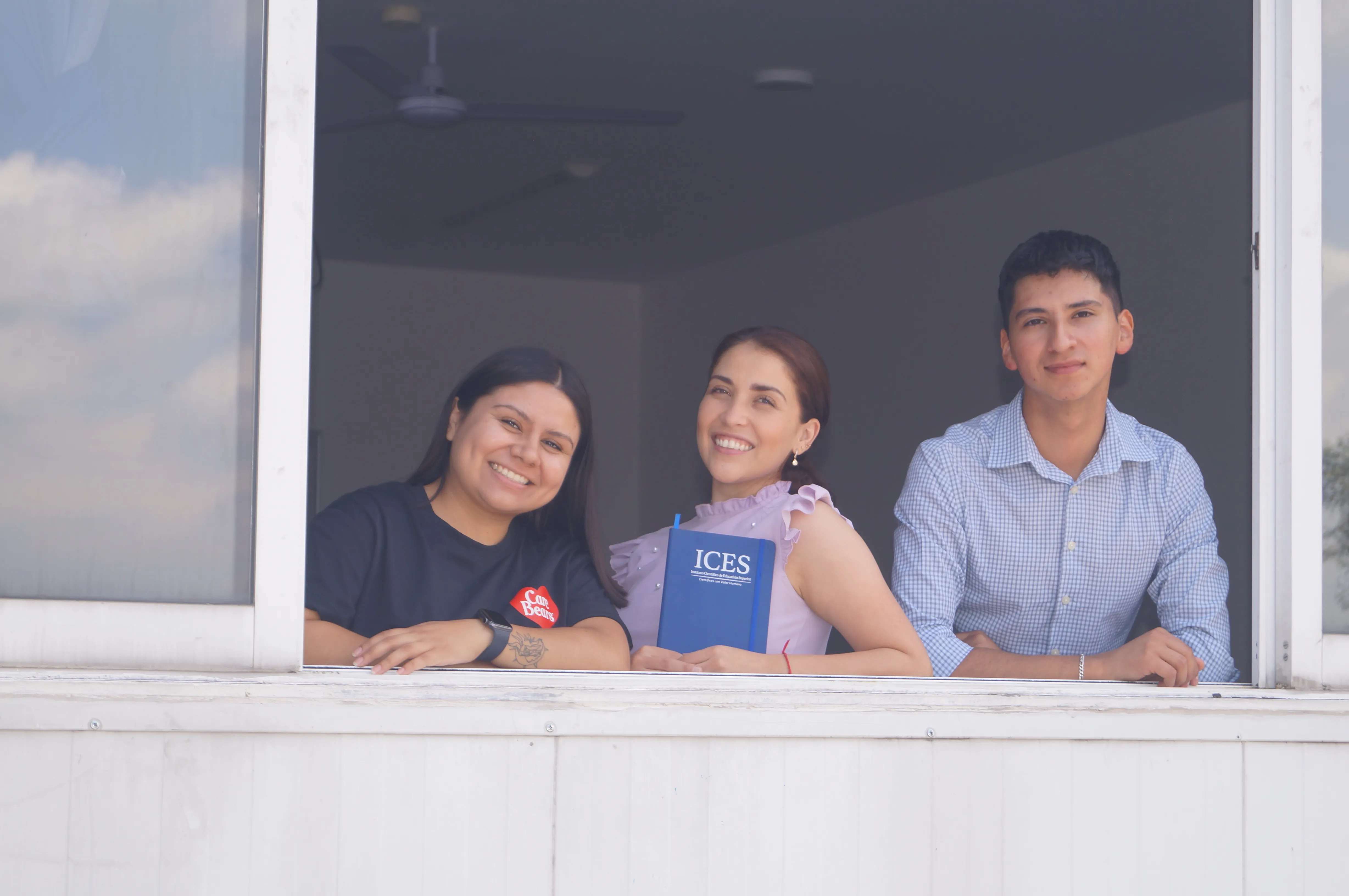Tres estudiantes de ICES asomados a la ventana de un aula, una con una libreta institucional